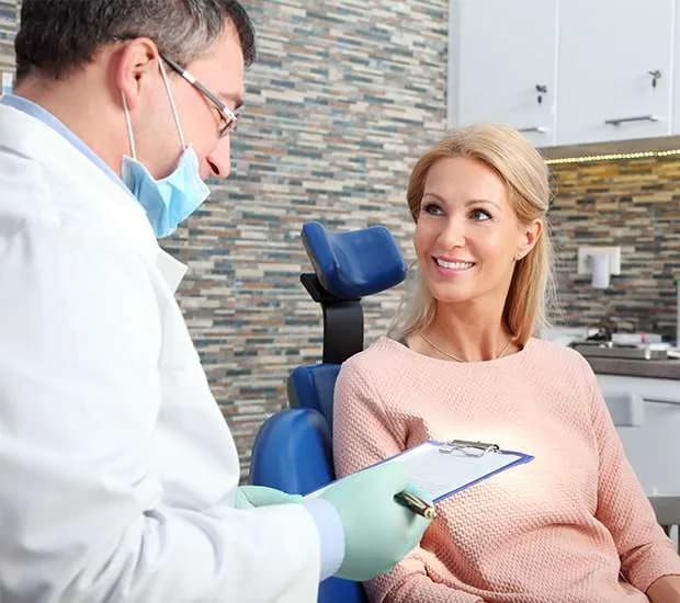 A dentist in a mask holding a clipboard talks with a smiling woman seated in a dental chair.