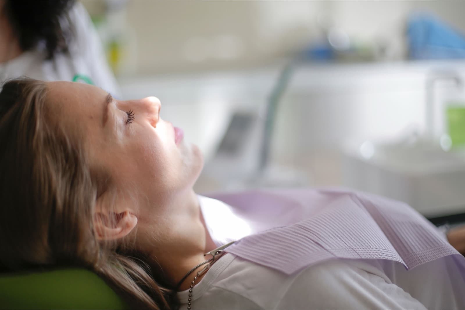 A woman in profile reclined in a dental chair, a paper bib clipped at her neck and her eyes closed.