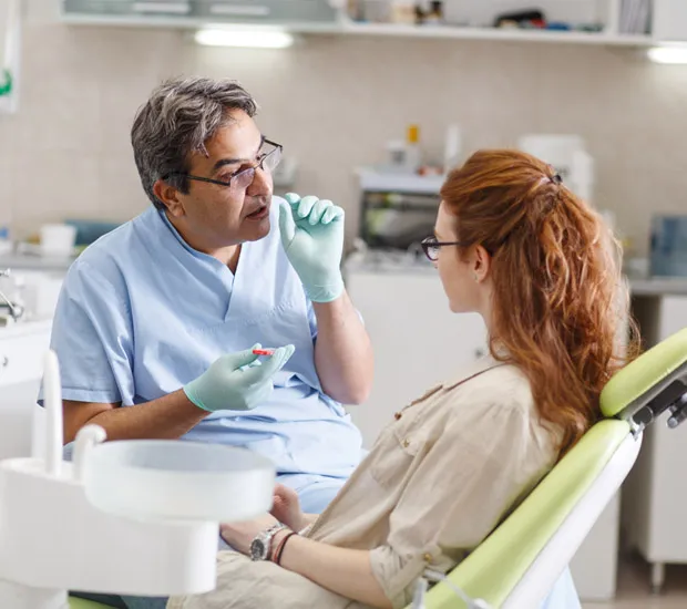 A dentist in scrubs talks with a woman seated in a dental chair.
