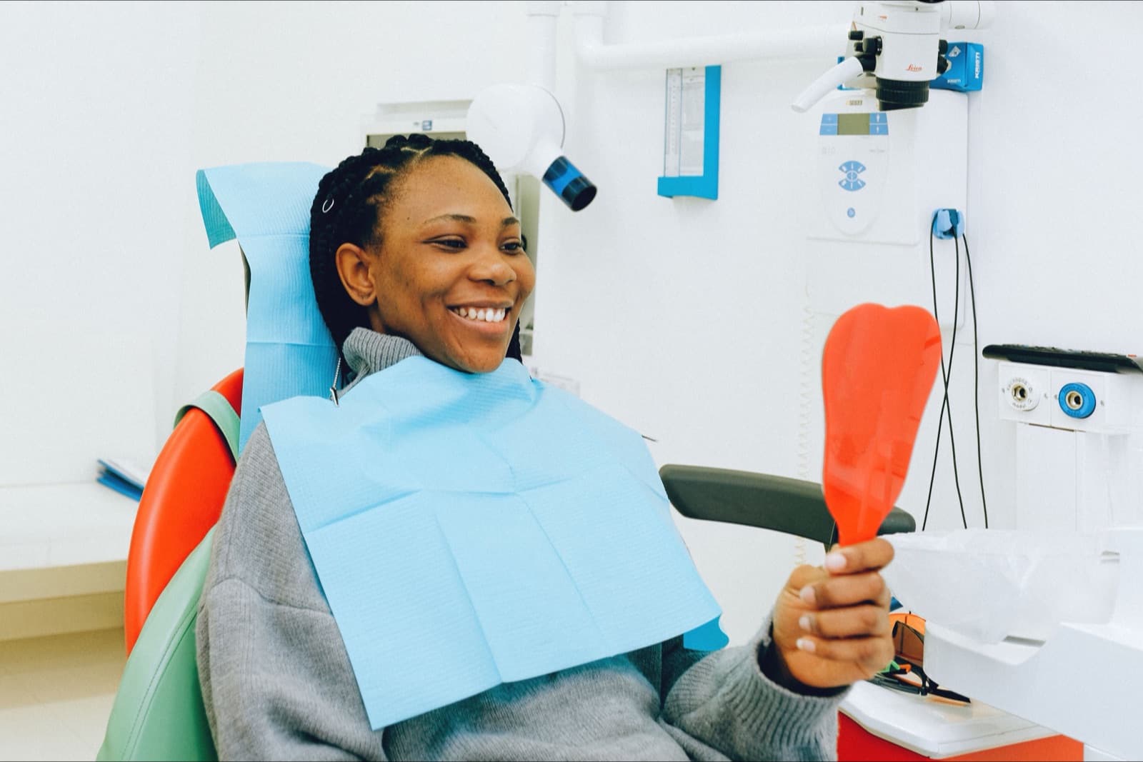 A woman in a dental chair wearing a blue bib holds a hand mirror up to her teeth.