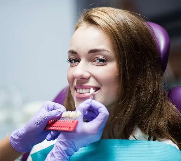 A smiling woman in a dental chair as a dental professional holds a tooth shade guide beside her teeth.
