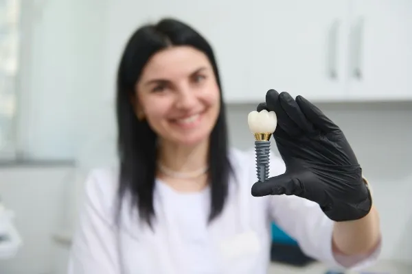 A smiling woman in a white coat holds up a dental implant crown in a black-gloved hand.