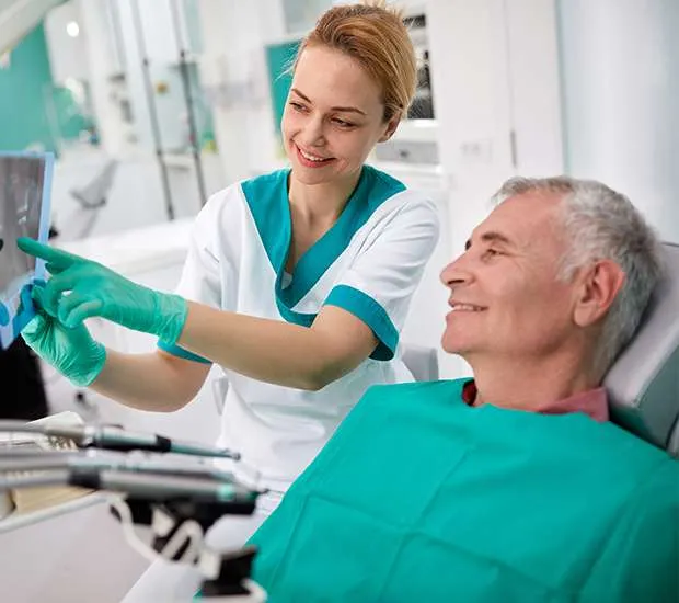 A dental assistant points at an x-ray while an older man sits in the dental chair.