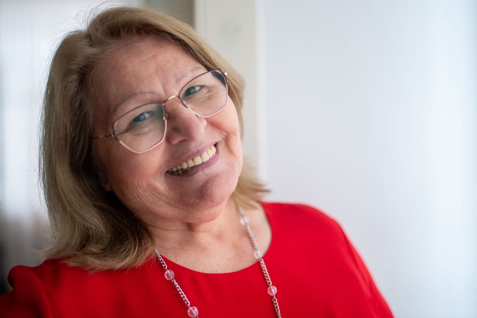 A smiling woman in her sixties with blonde hair, wire-rim glasses and a red top, photographed against a white wall in daylight.