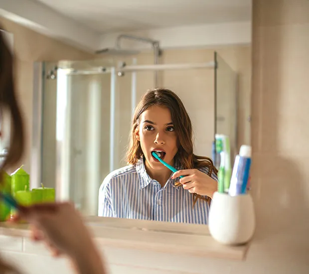 A woman brushing her teeth in a bathroom mirror, with toothpaste and brushes in a cup beside her.