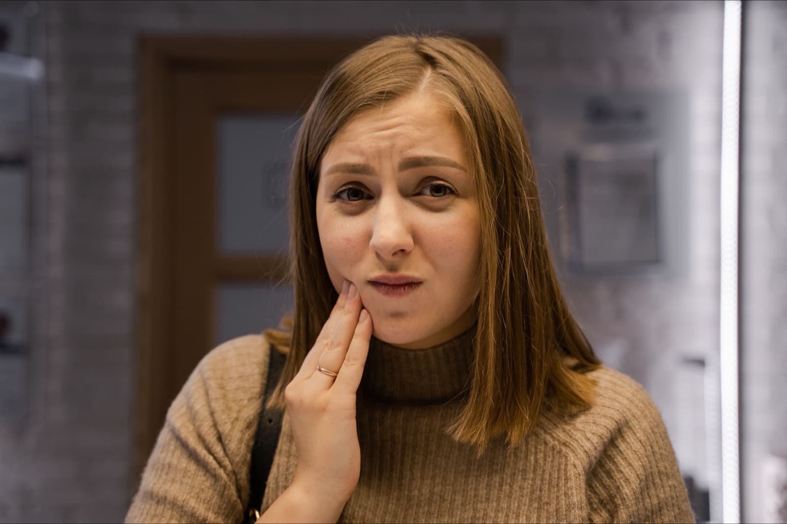 A woman in a tan knit sweater touches her cheek and jaw with a mild frown.
