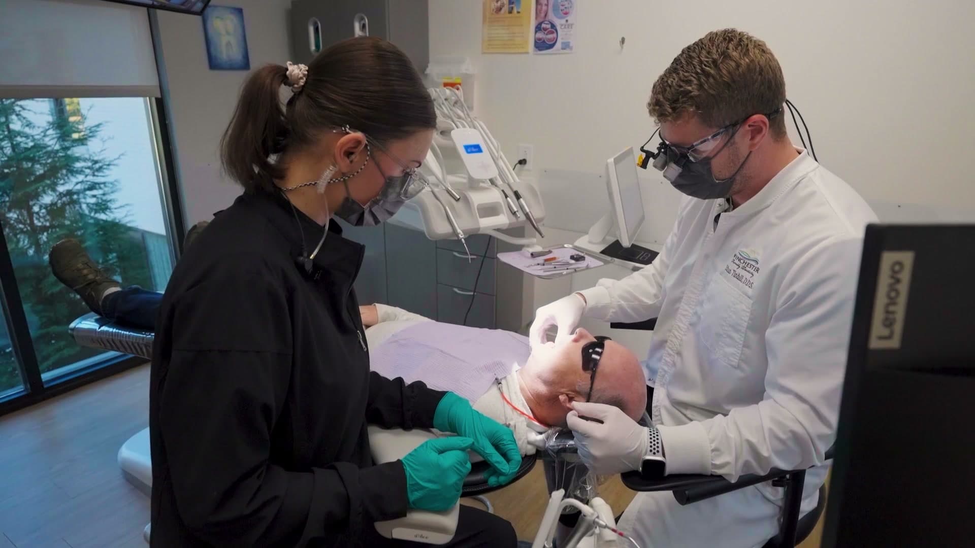 A dentist in a white coat and magnifying loupes and a dental assistant in black scrubs treating a patient reclined in the dental chair.