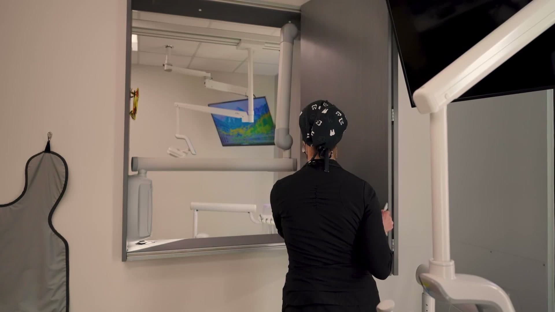 A staff member in black scrubs and a tooth-print cap, seen from behind, at an open wall cabinet beside a ceiling-mounted exam light and a wall-mounted screen.