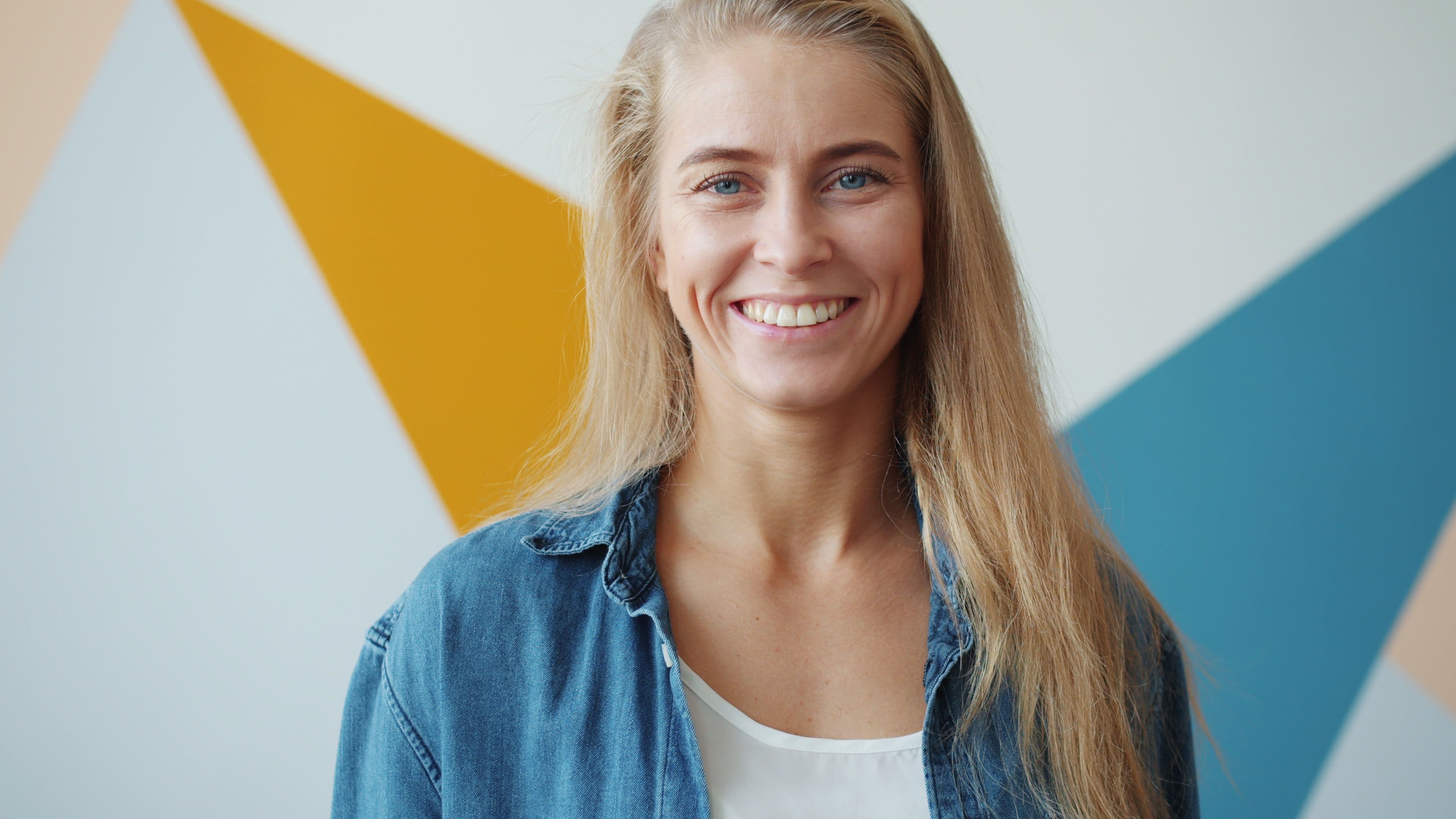 A smiling woman with long blonde hair and a denim shirt, in front of a painted wall of orange, blue and cream shapes.