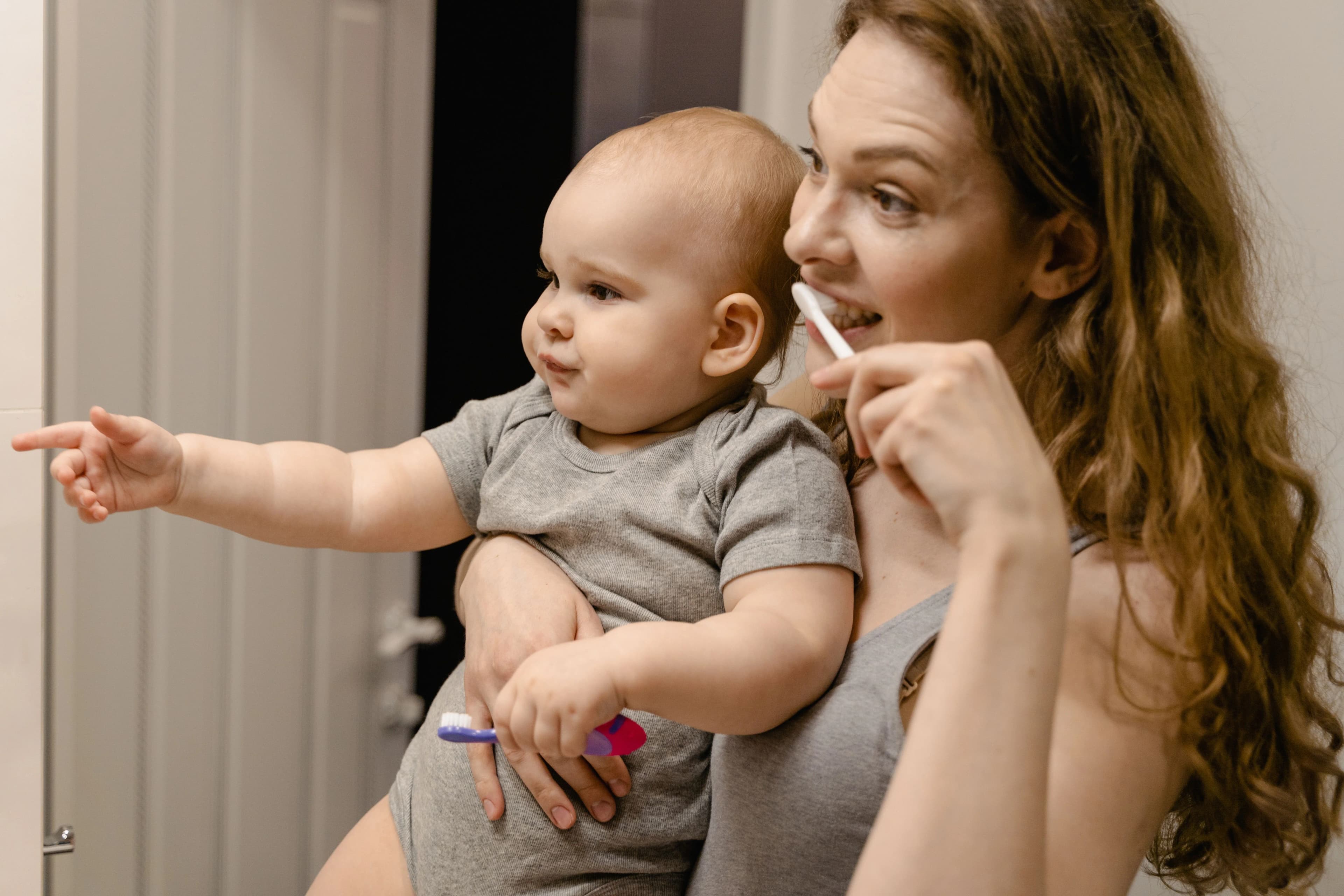A mother brushing her teeth while holding her baby, who reaches out towards the camera.