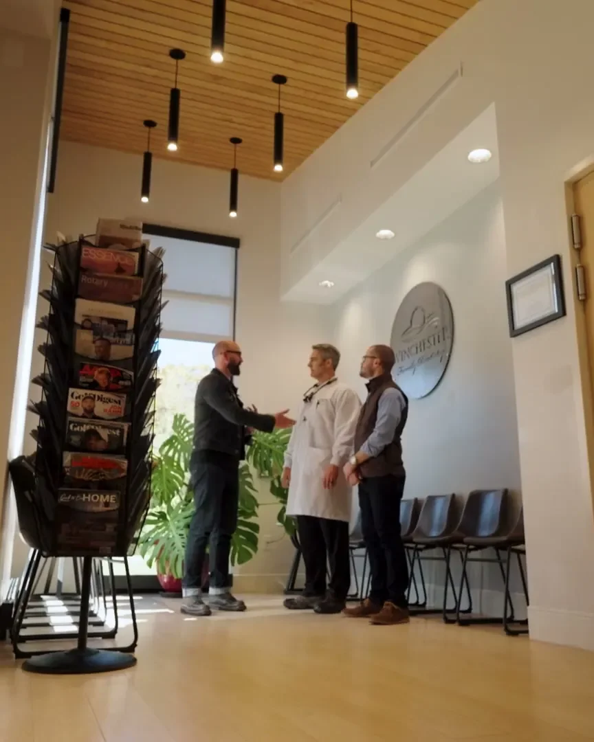 Two men and a dentist in a white coat shaking hands in the practice's front lobby, with the brochure rack beside them.