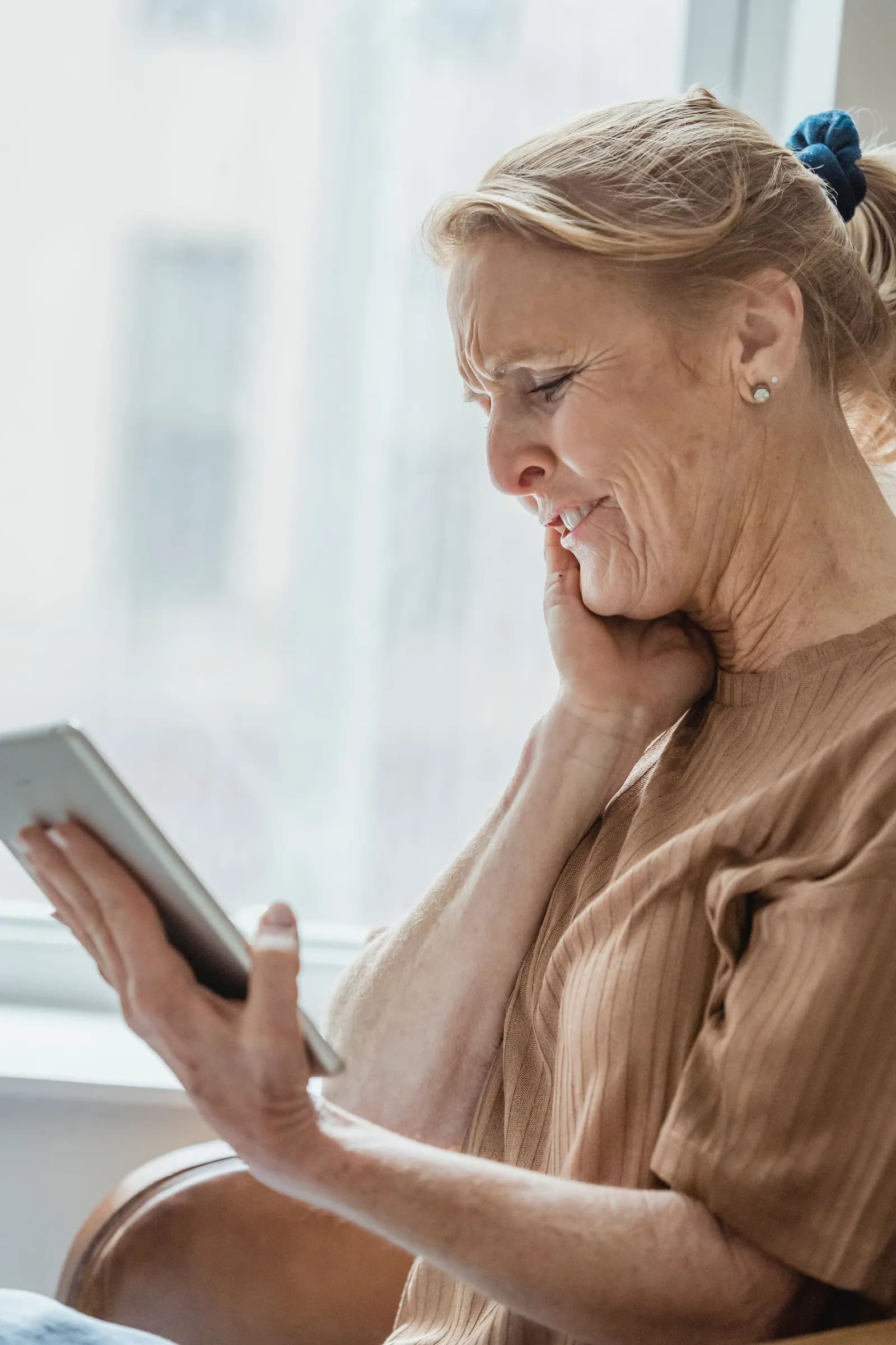 A woman with her hand against the side of her jaw, wincing, as she looks at a tablet.