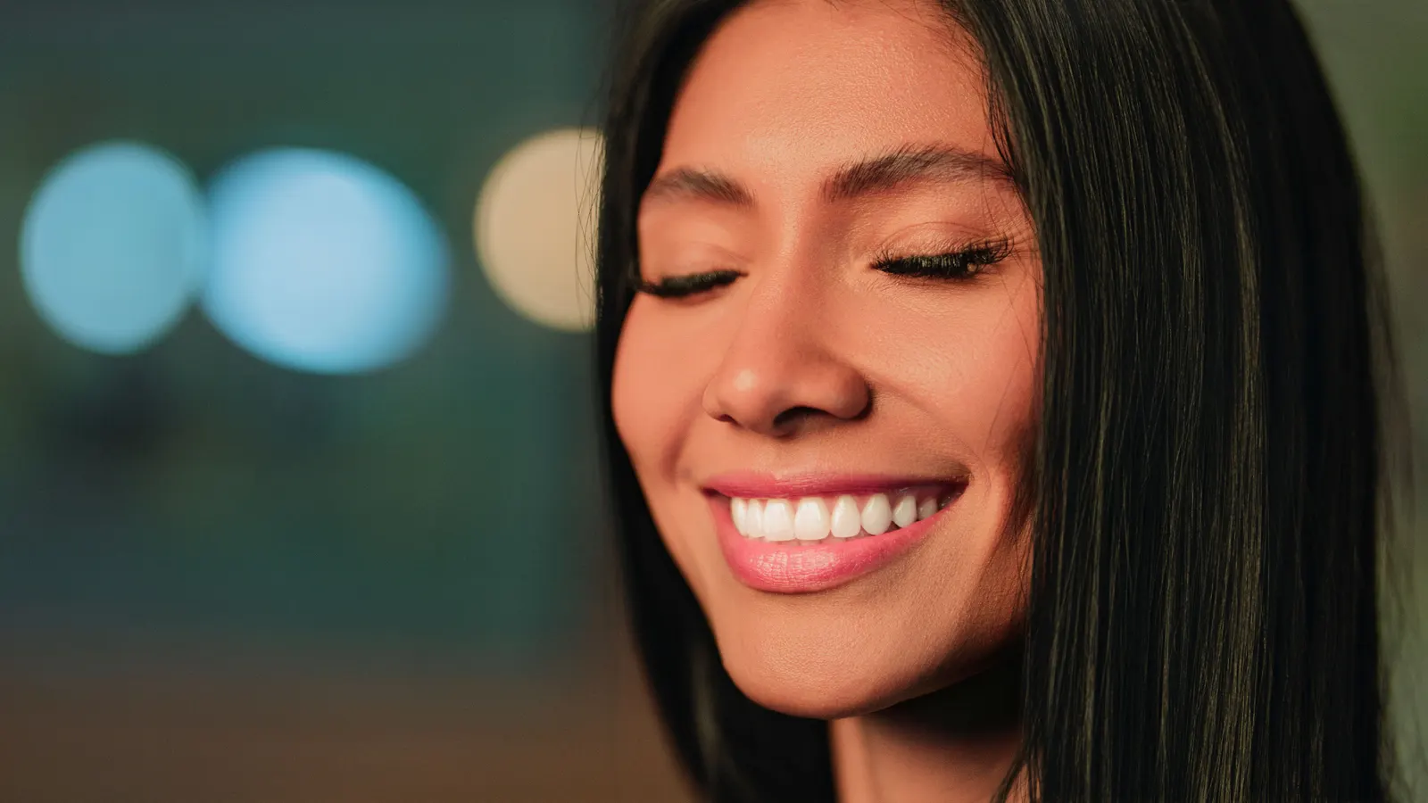 A woman smiling with her eyes closed, photographed close against a dark background.