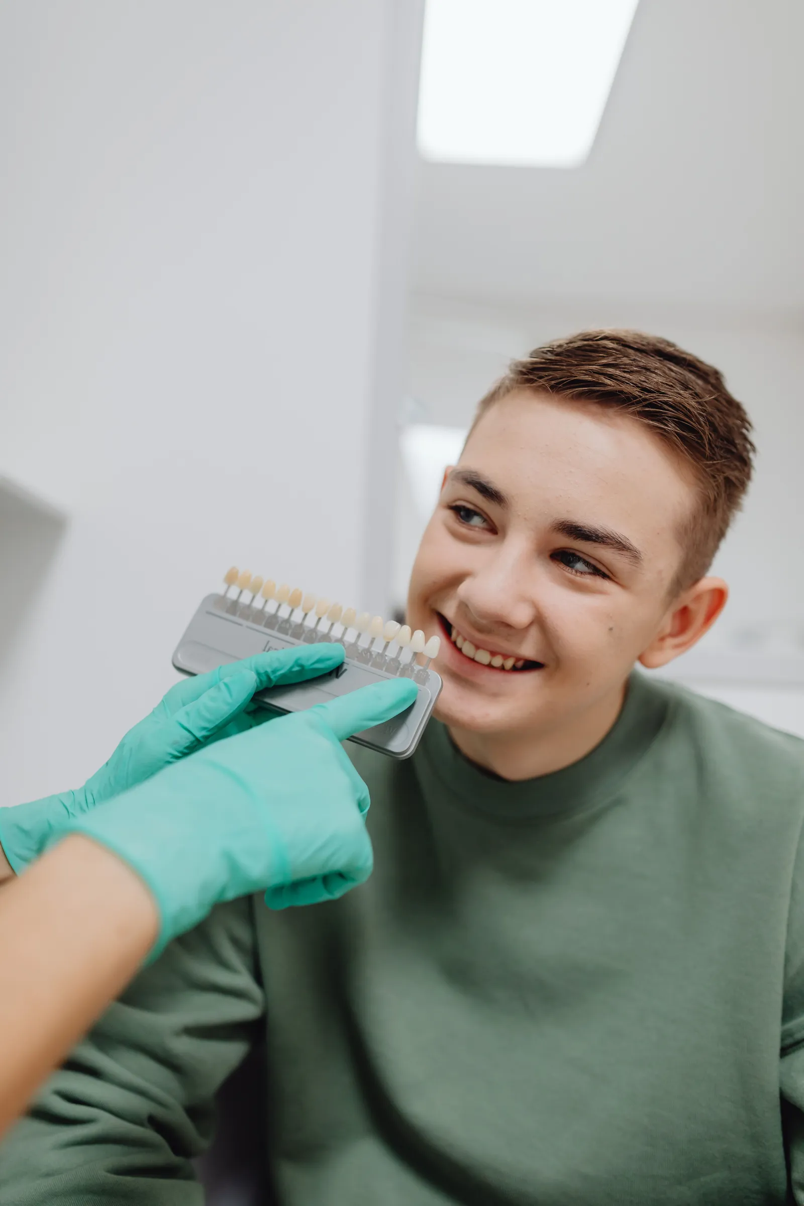 A smiling teenager in a green sweater as a gloved hand holds a tooth shade guide beside his teeth.