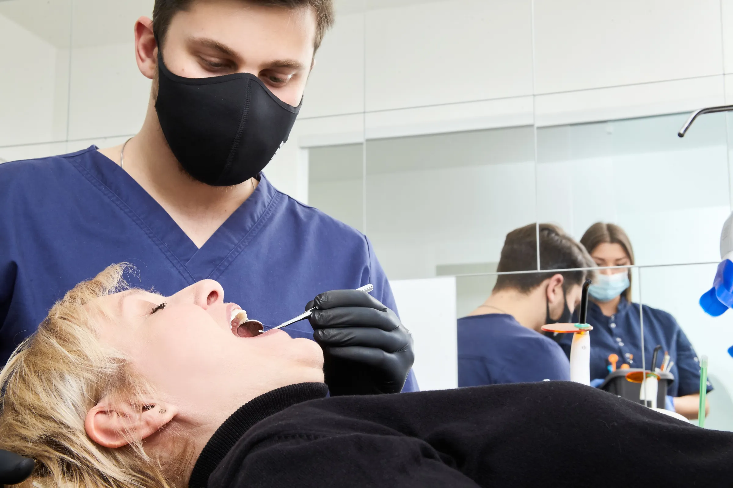 A patient reclined in a dental chair while a dentist in a mask and blue scrubs examines her mouth with a mirror.