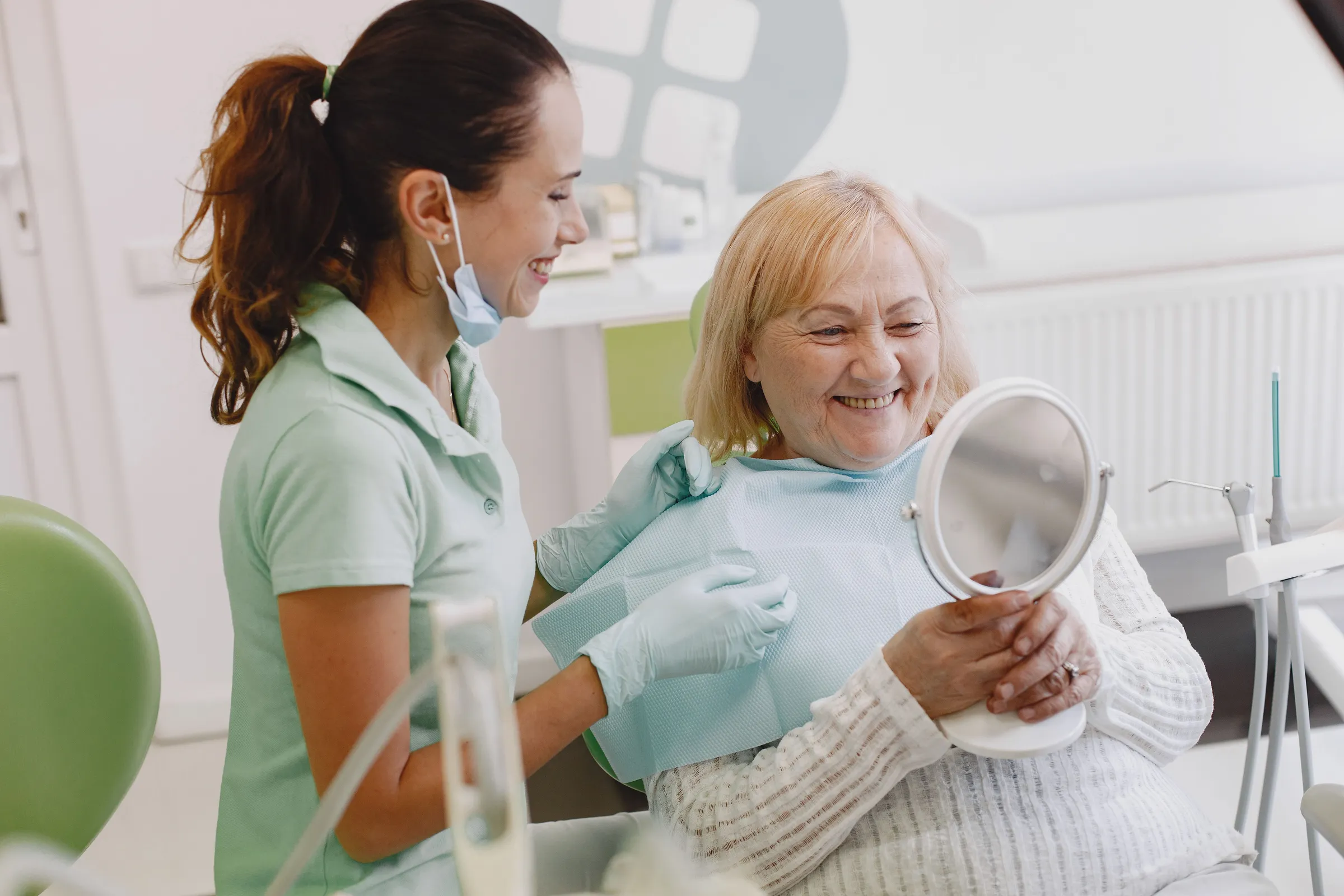 An older woman smiling at her reflection in a hand mirror while a hygienist stands beside her.