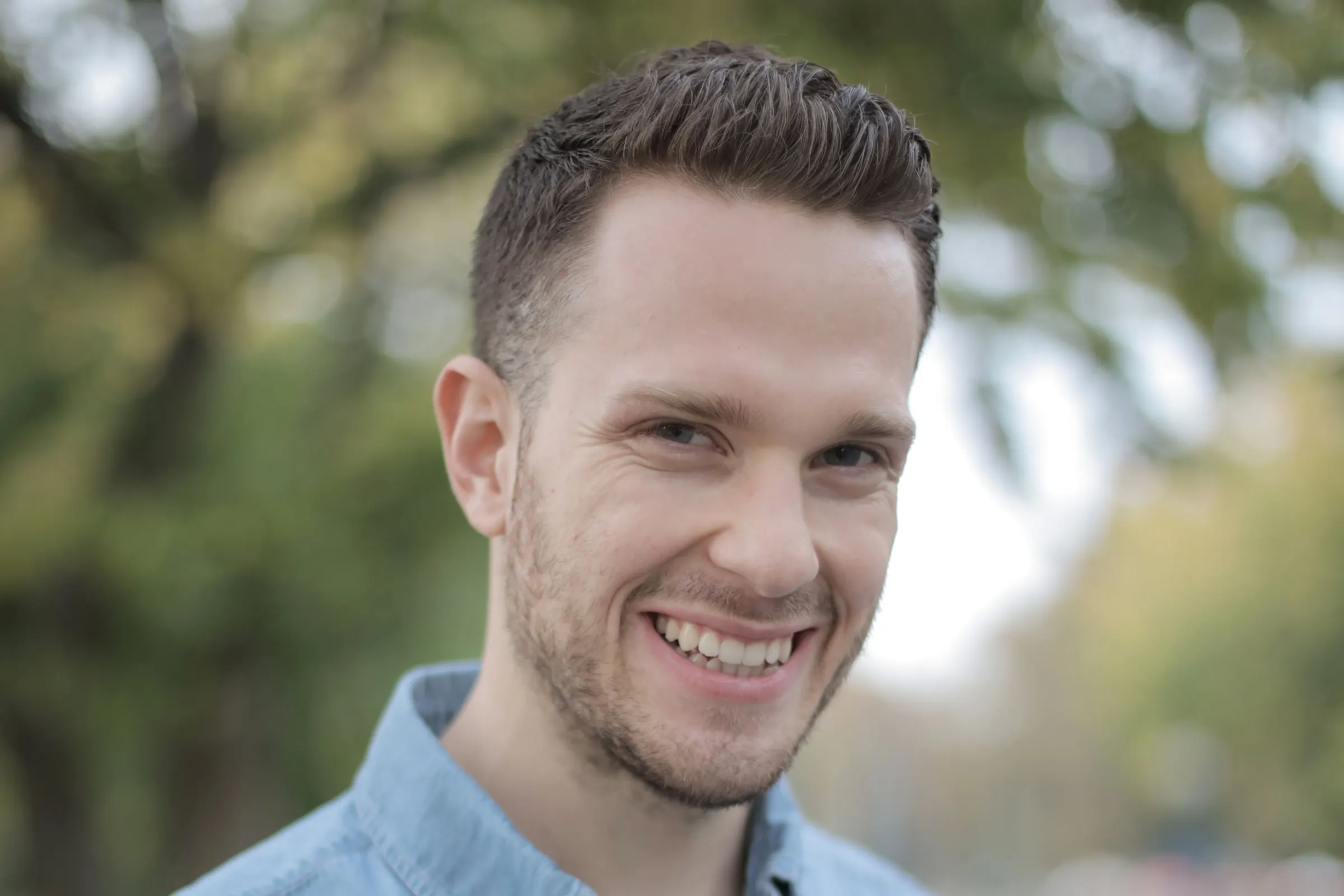 A man in a light-blue shirt smiling outdoors, trees blurred behind him.