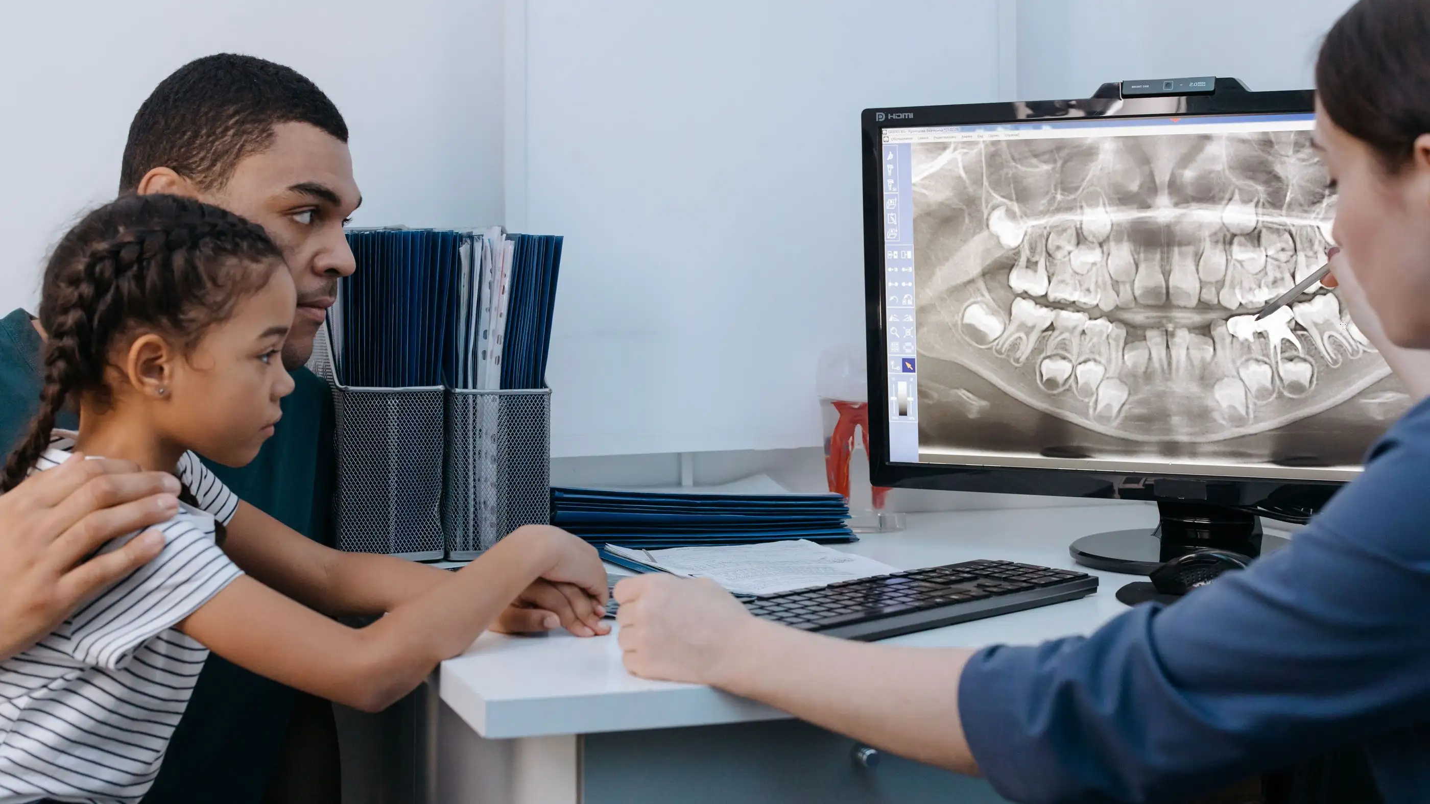 A young girl and her father watching a dental hygienist point out her X-ray on a computer screen.