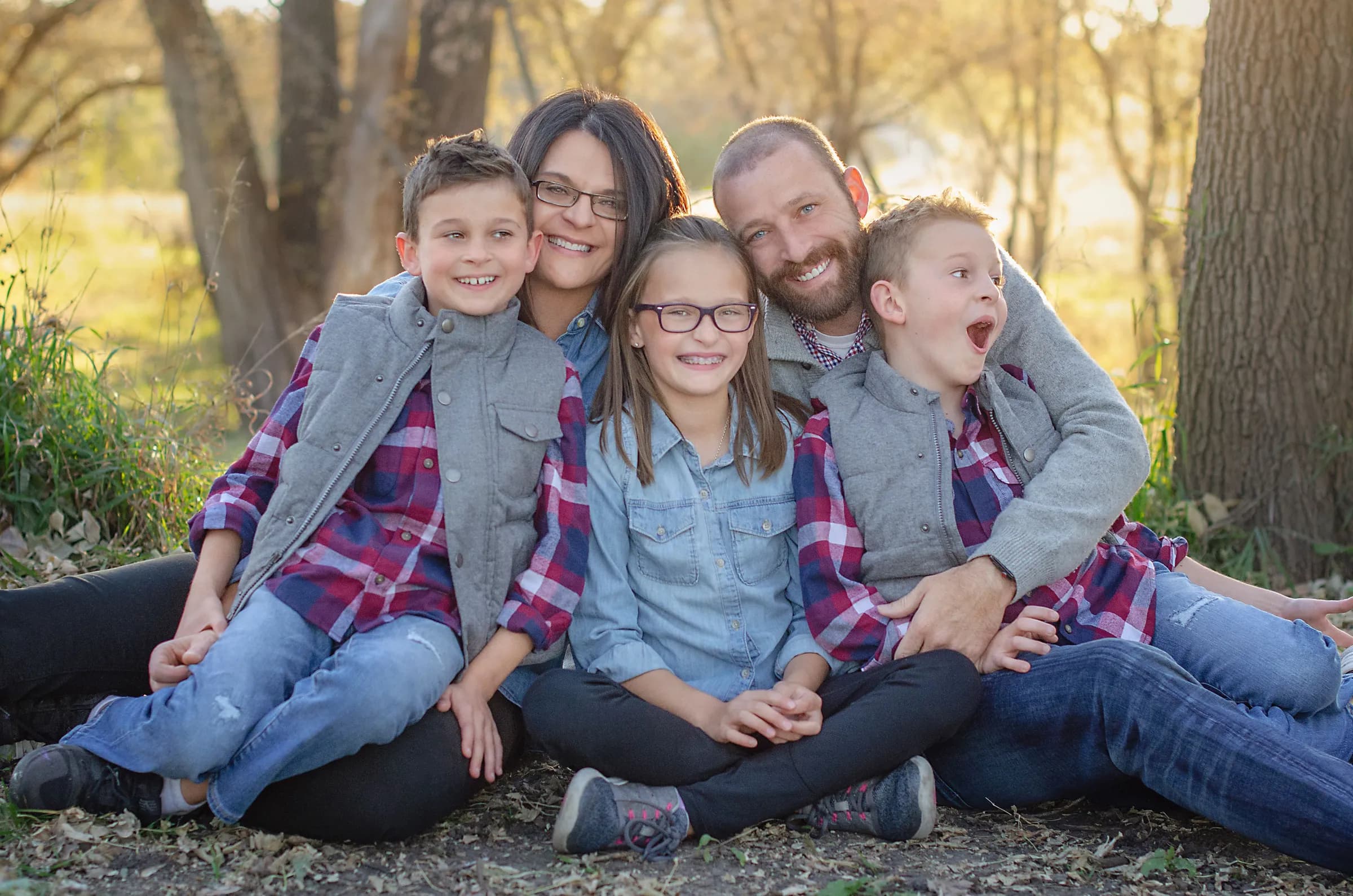 A mother, a father and their three children sitting close together on the ground outdoors, all smiling.