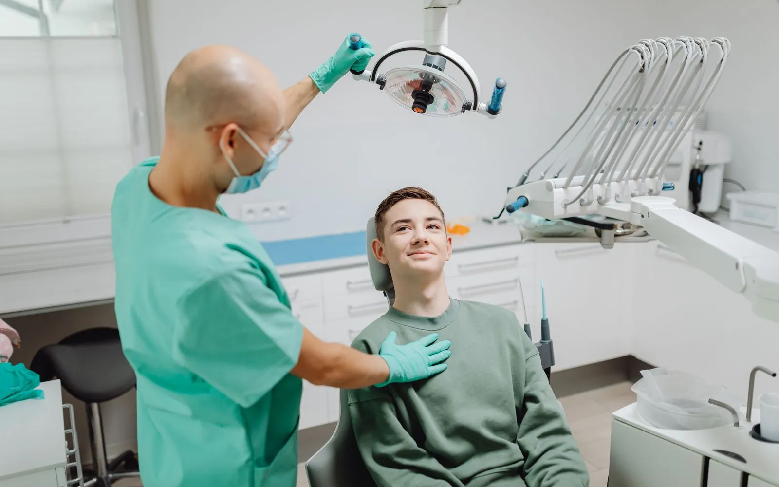 A teenager in a green sweater sitting in a dental chair, smiling up at a dentist in green scrubs who stands beside him.
