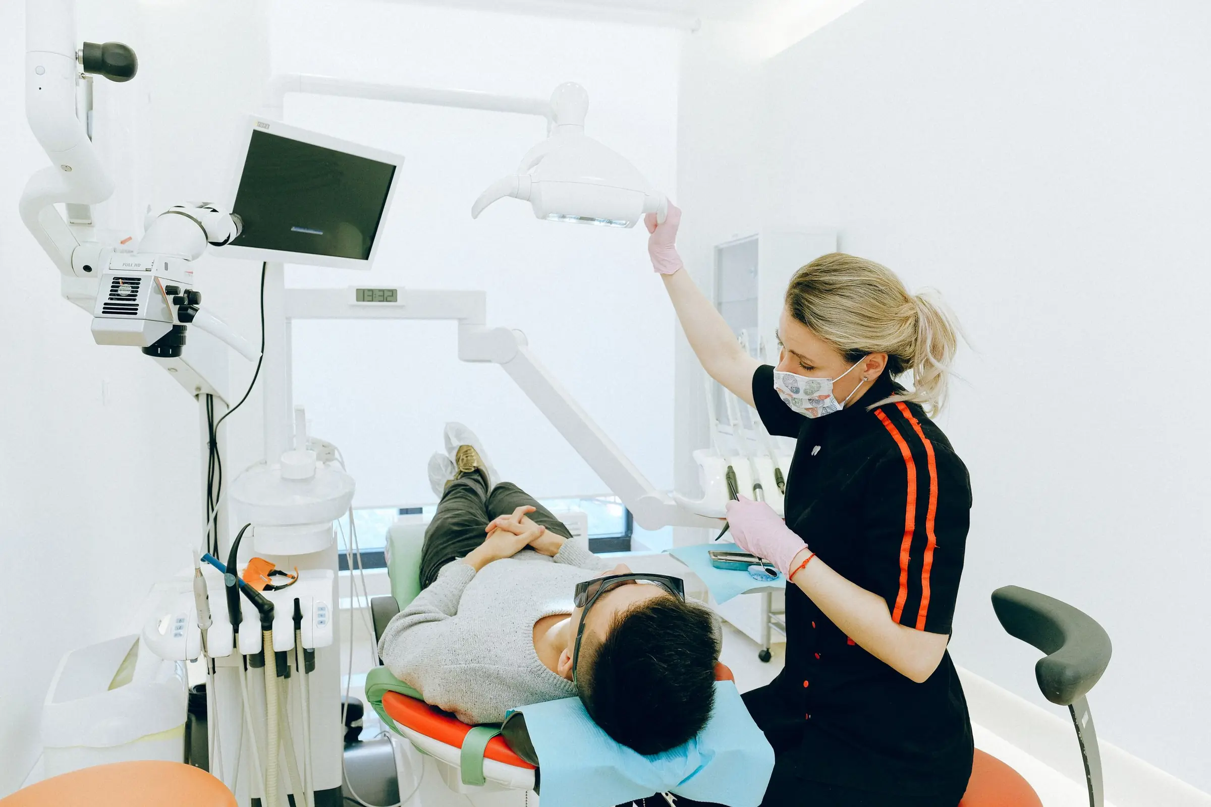 A dental hygienist in a black uniform with a patterned face mask adjusts the overhead light while a patient in a grey sweater lies back in the chair of a bright, white treatment room.