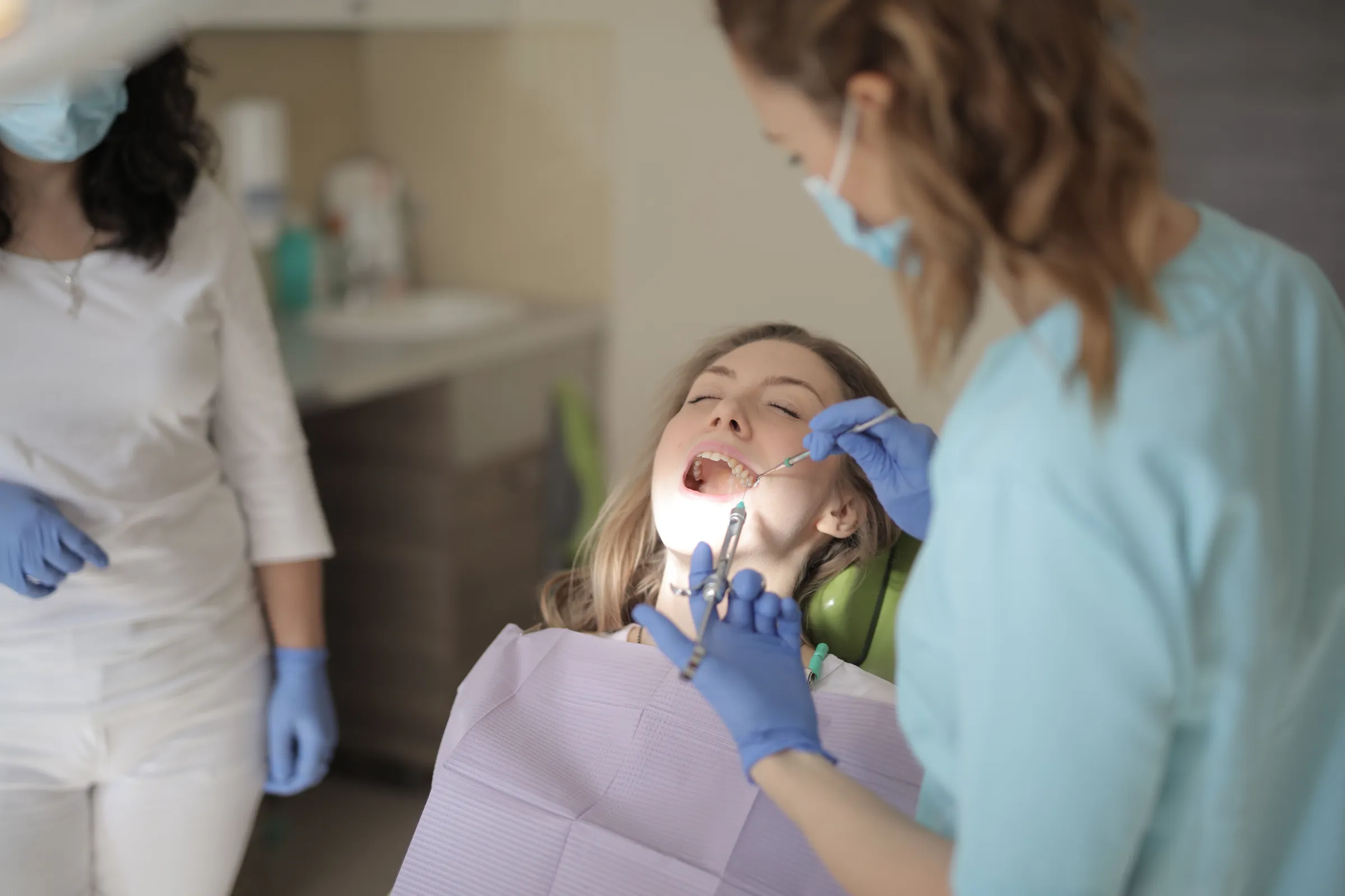 A young woman in a dental chair having her teeth cleaned, with a hygienist working and a second person standing by.