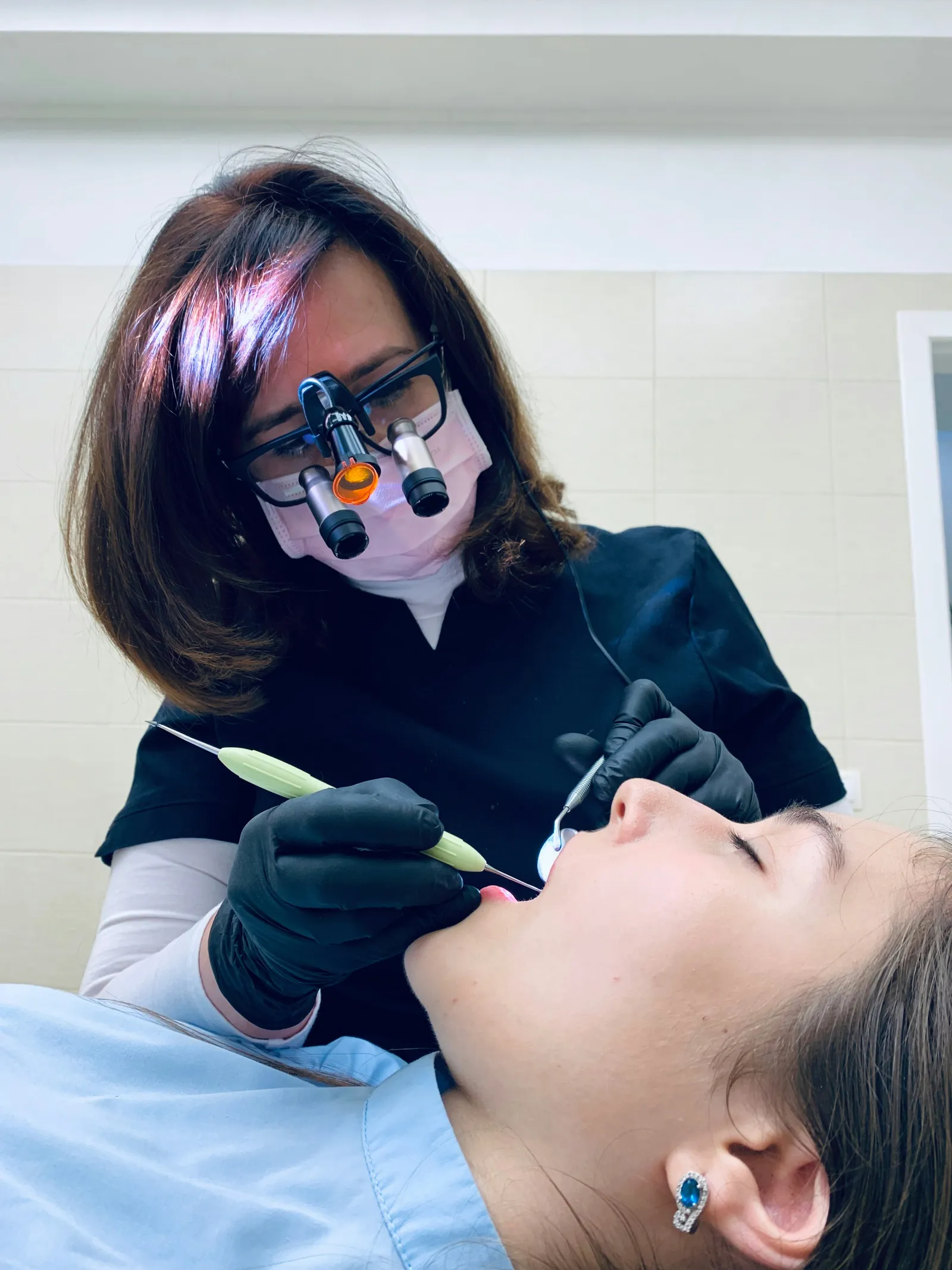 A clinician in magnifying loupes and a headlight works at a reclined patient's open mouth with a mirror and an instrument.