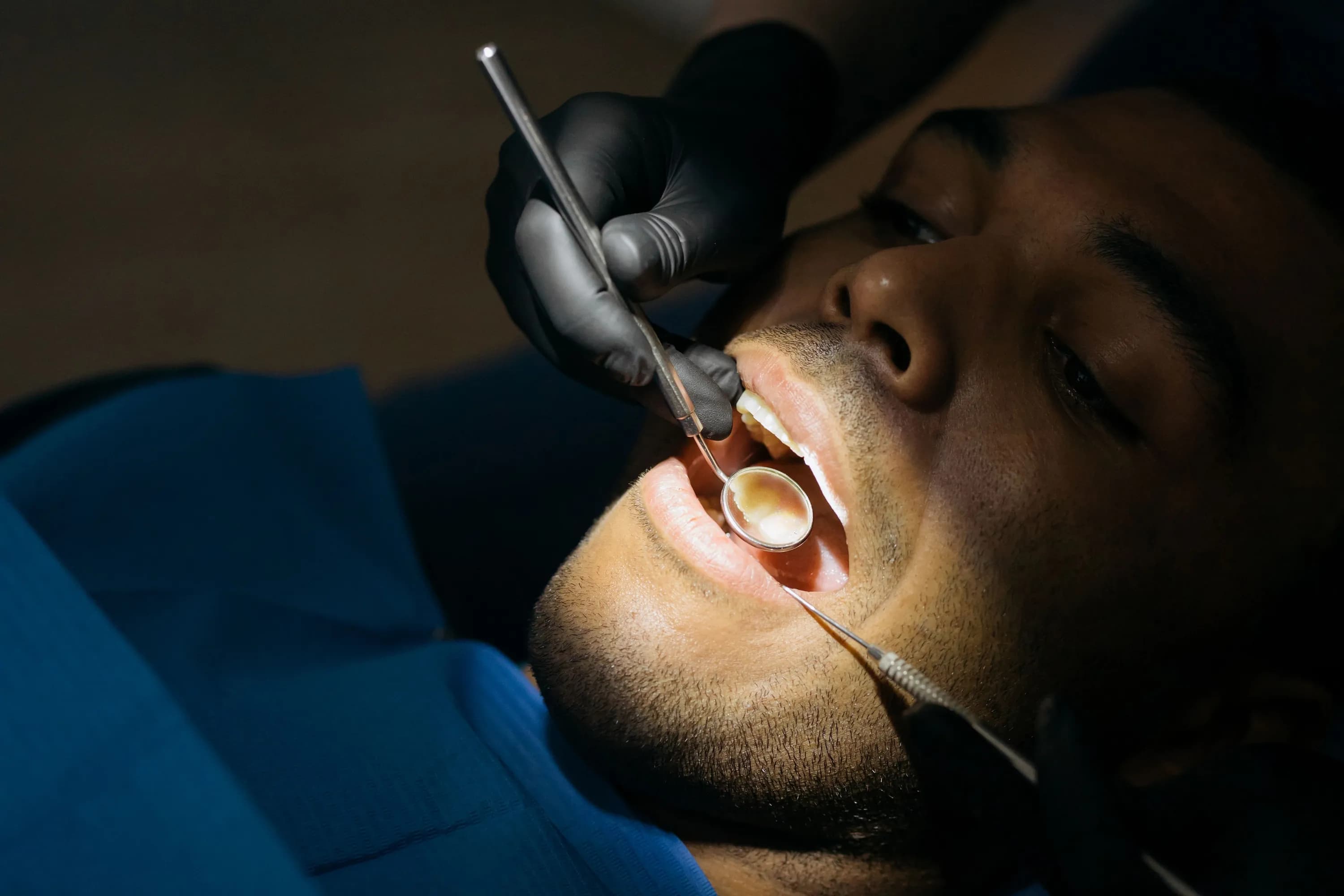 A dentist's gloved hands hold a mouth mirror and a probe to a man's teeth and gums as he lies back in the chair.
