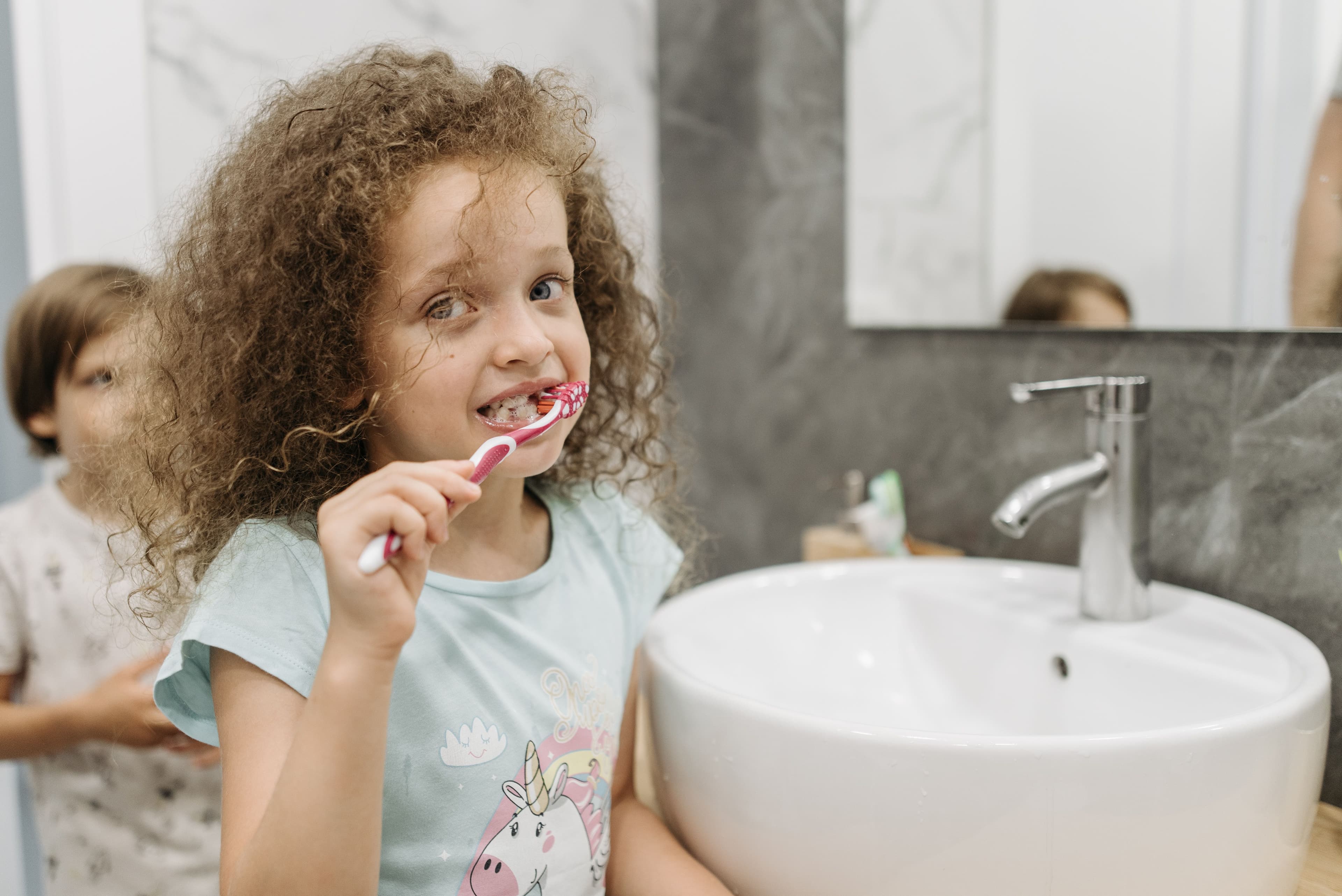 A young girl with curly hair brushing her teeth at a bathroom sink.