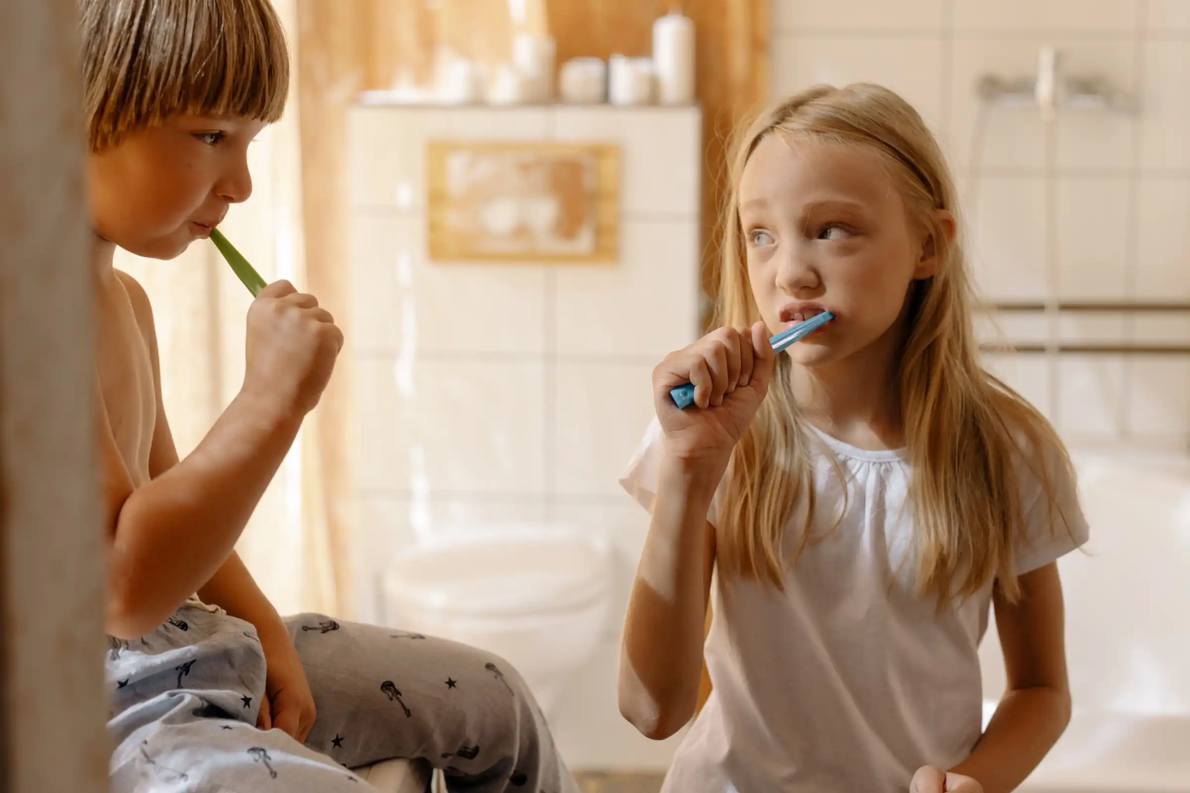 A girl brushing her teeth in a sunlit bathroom, her brother beside her.