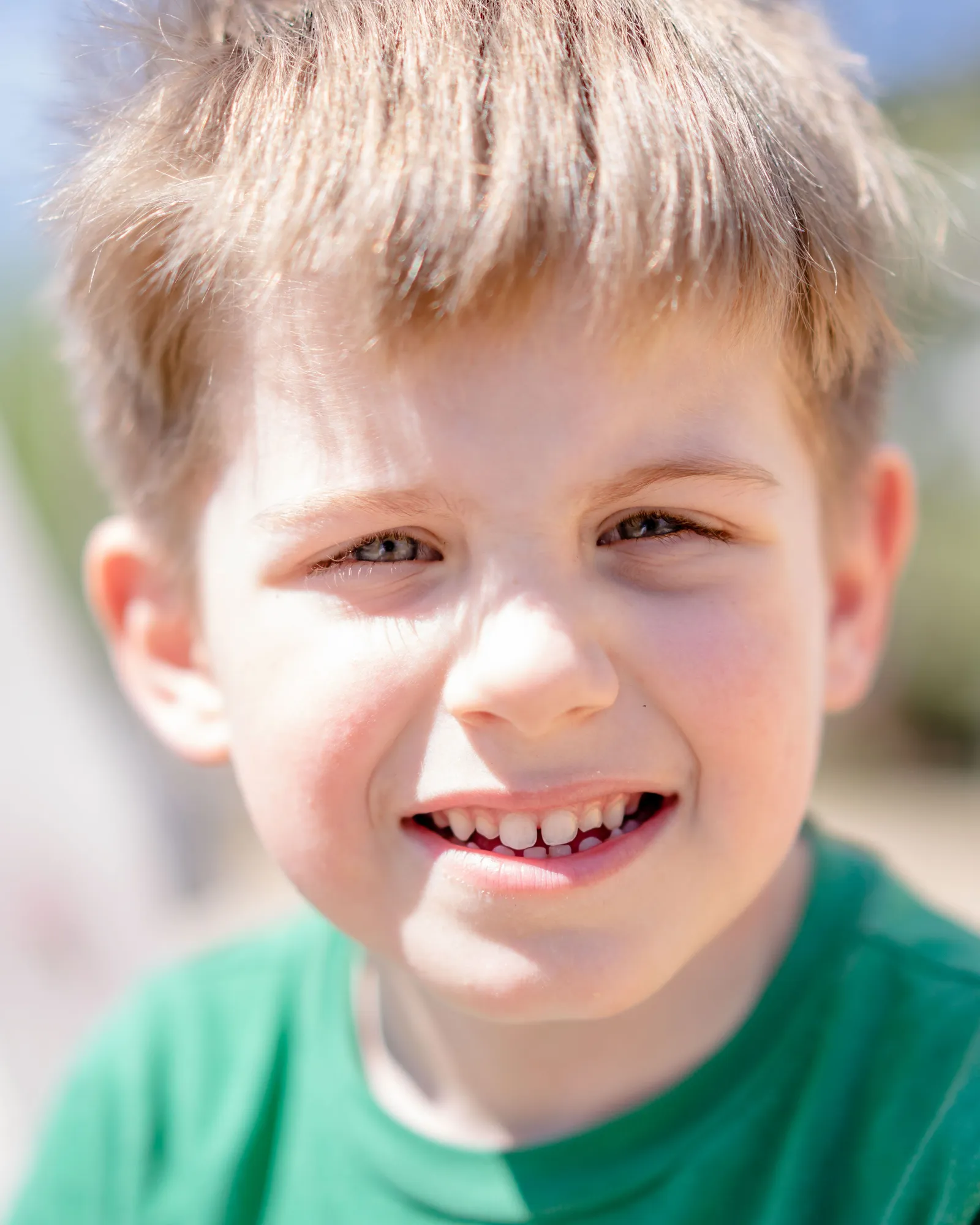 A boy in a green T-shirt smiling at the camera, outdoors.