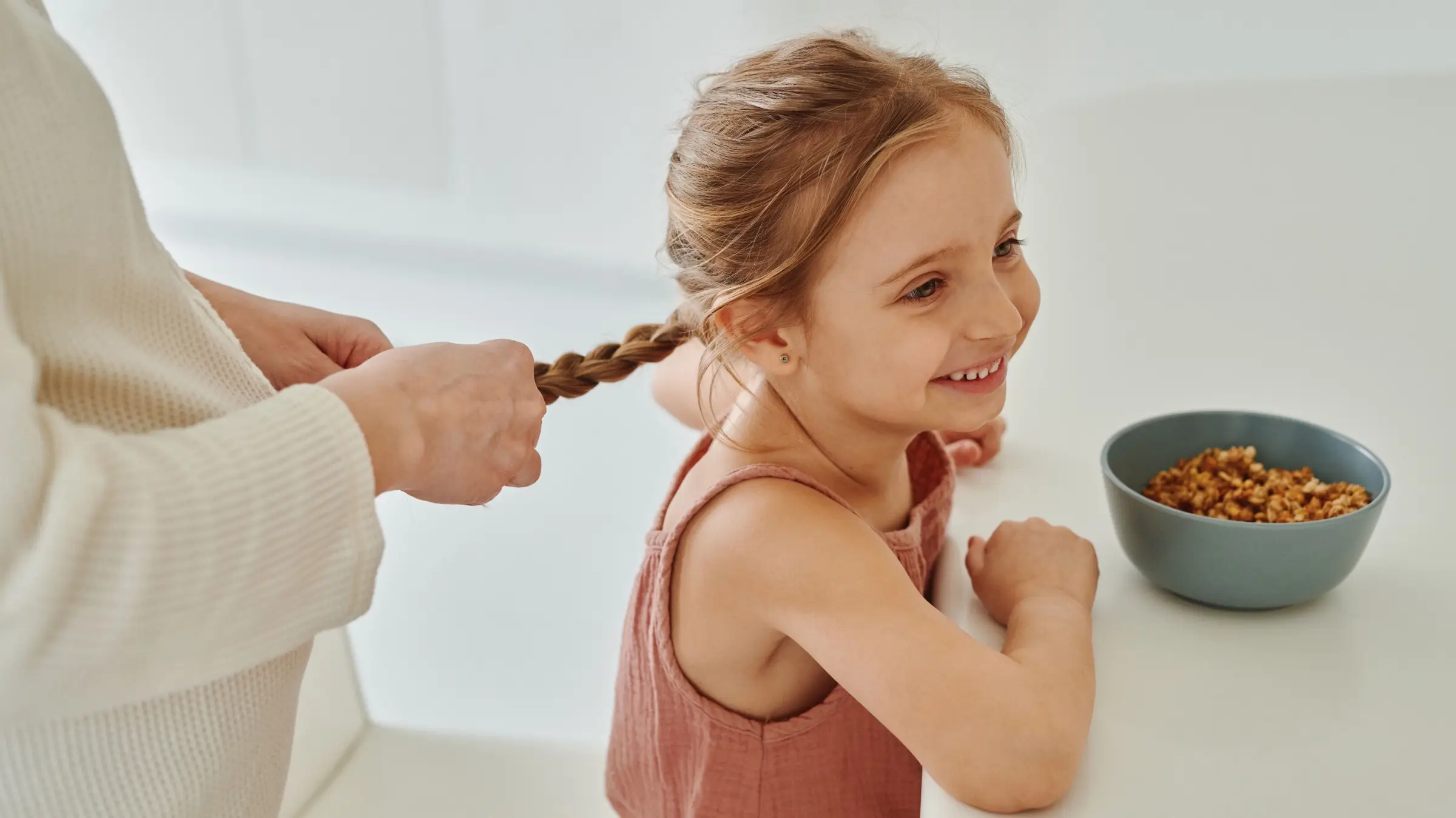 A young girl smiling at a kitchen table beside a bowl of cereal while her mother braids her hair.