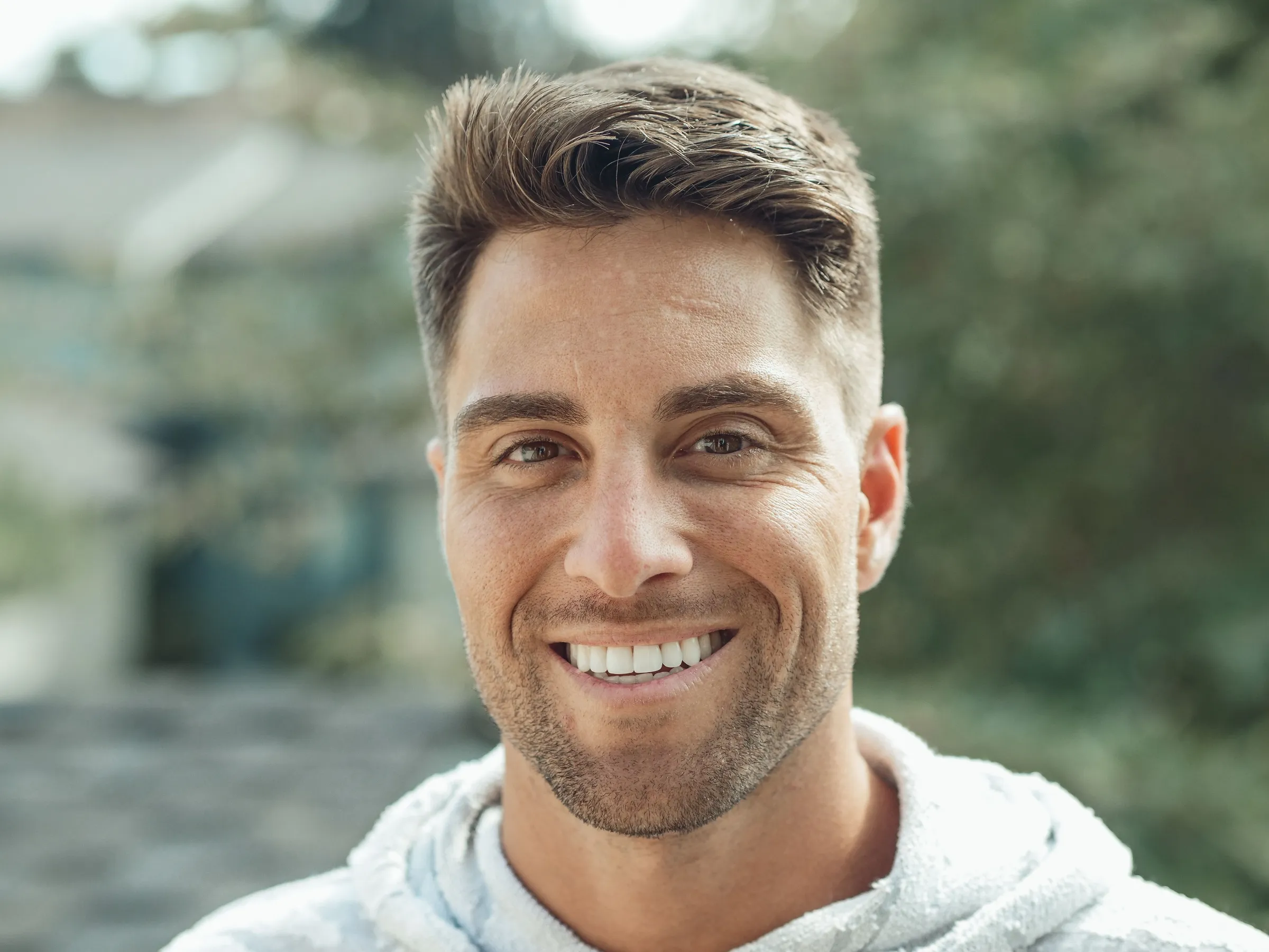 A man in a light grey hoodie smiling at the camera outdoors.