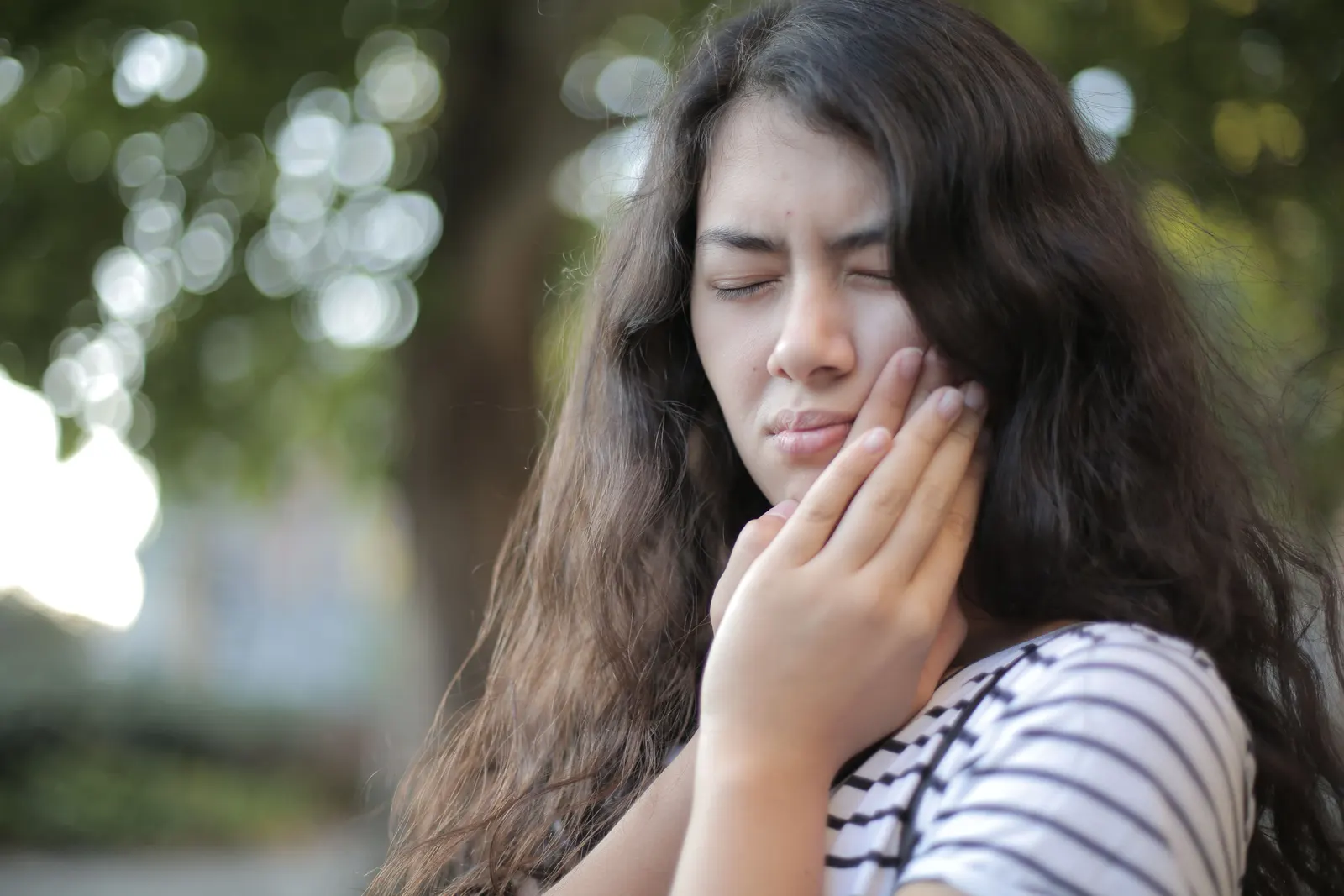 A woman outdoors with her hand held against her jaw, eyes closed.