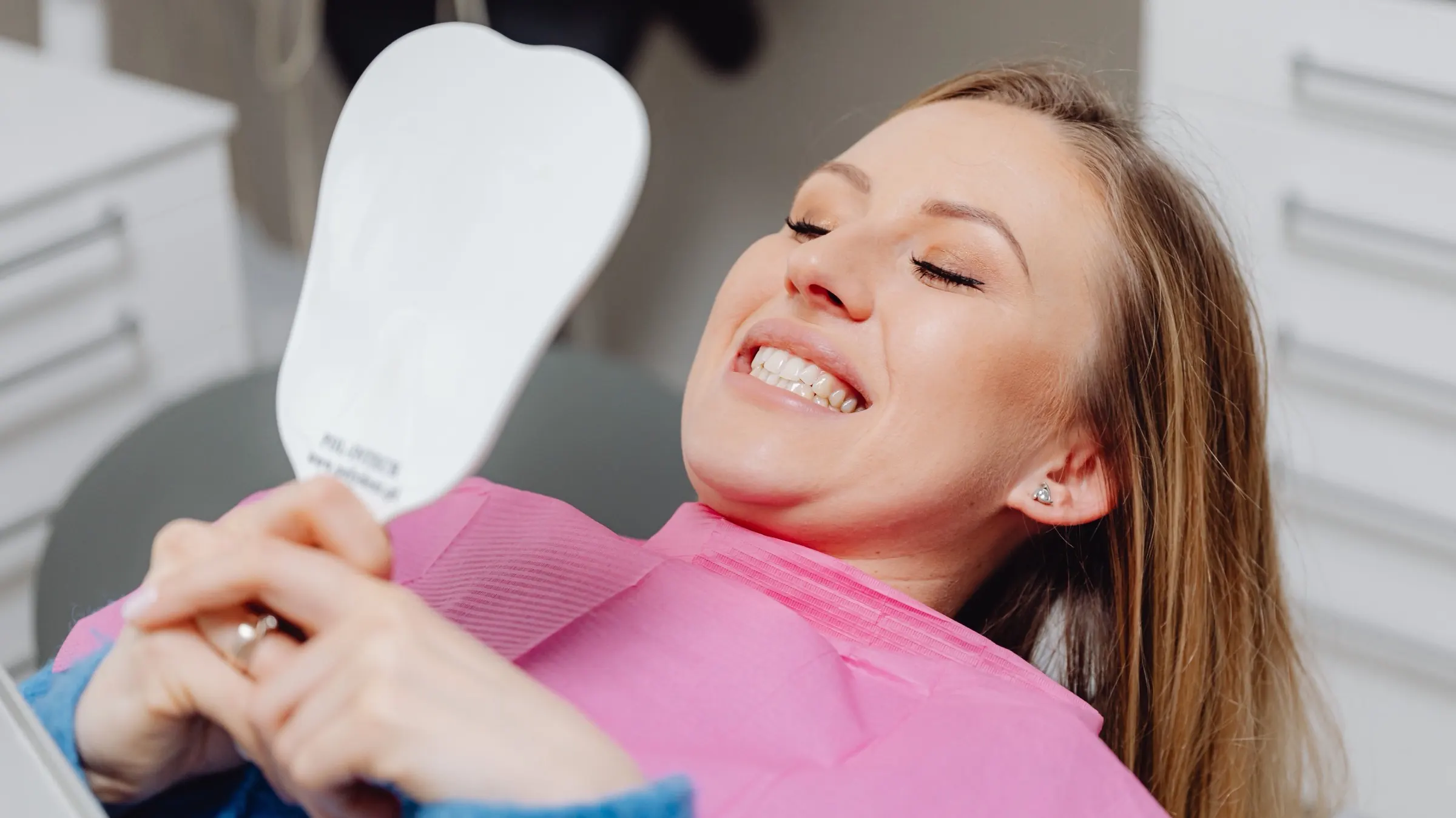 A woman reclining in a dental chair, smiling broadly with her eyes closed while holding a hand mirror.