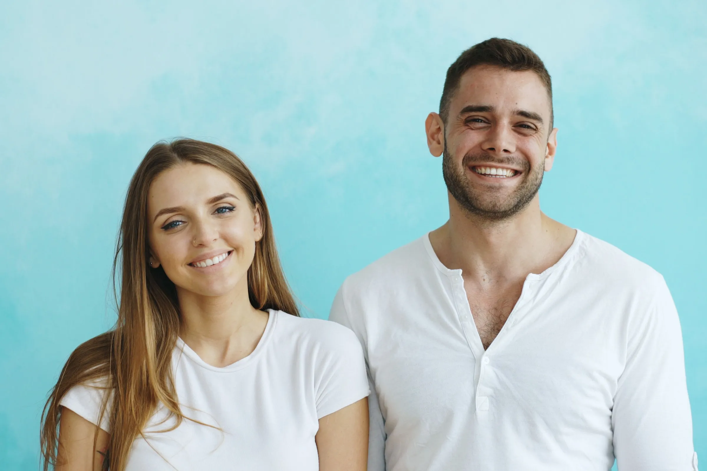A woman and a man standing side by side against a teal wall, both smiling at the camera.